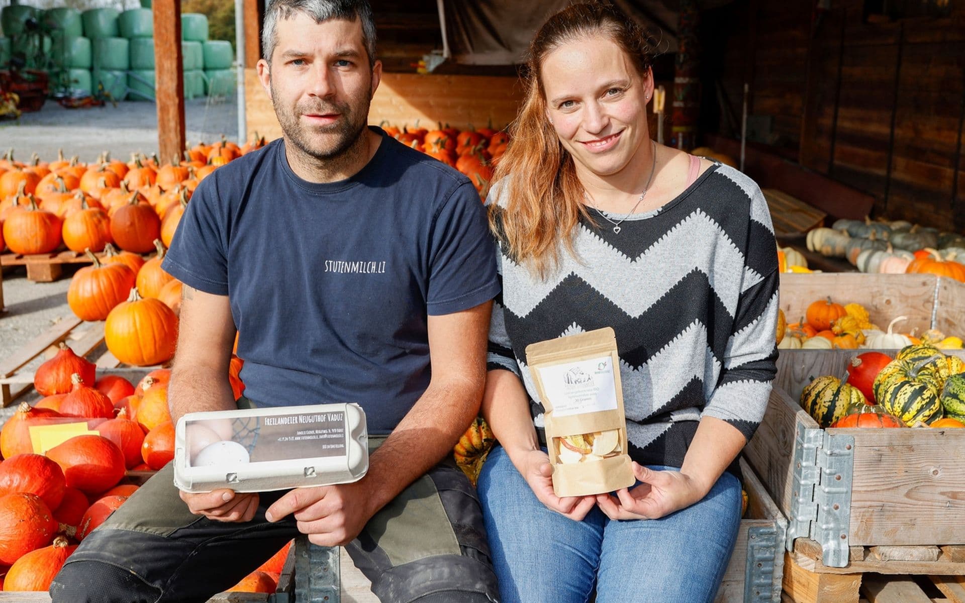Ein Mann und eine Frau sitzen zwischen Kürbissen und halten verpackte Waren in der Hand. Der Schauplatz ist im Freien, wo Holzkisten und verschiedene Kürbisse zu sehen sind.