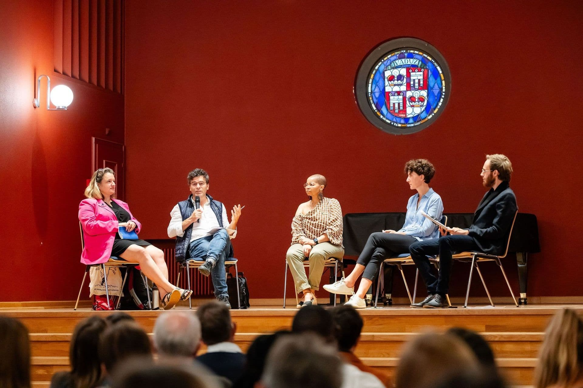 Fünf Personen sitzen bei einer Podiumsdiskussion auf einer Bühne im Rathaussaal, im Hintergrund ist ein Buntglasfenster zu sehen. Im Vordergrund sind die Zuhörer zu sehen.