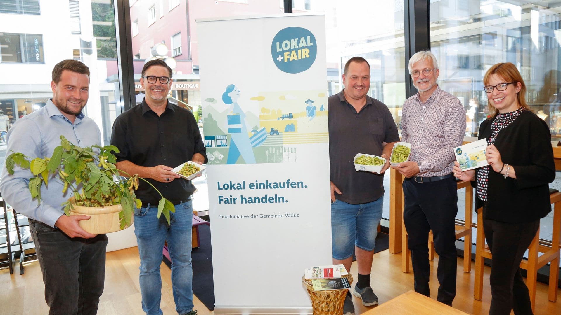 Fünf Personen stehen im Cafe im Kunstmuseum und präsentieren die erste Edamame Ernte aus Vaduz. Daneben steht ein Banner mit der Aufschrift „Lokal + Fair“.