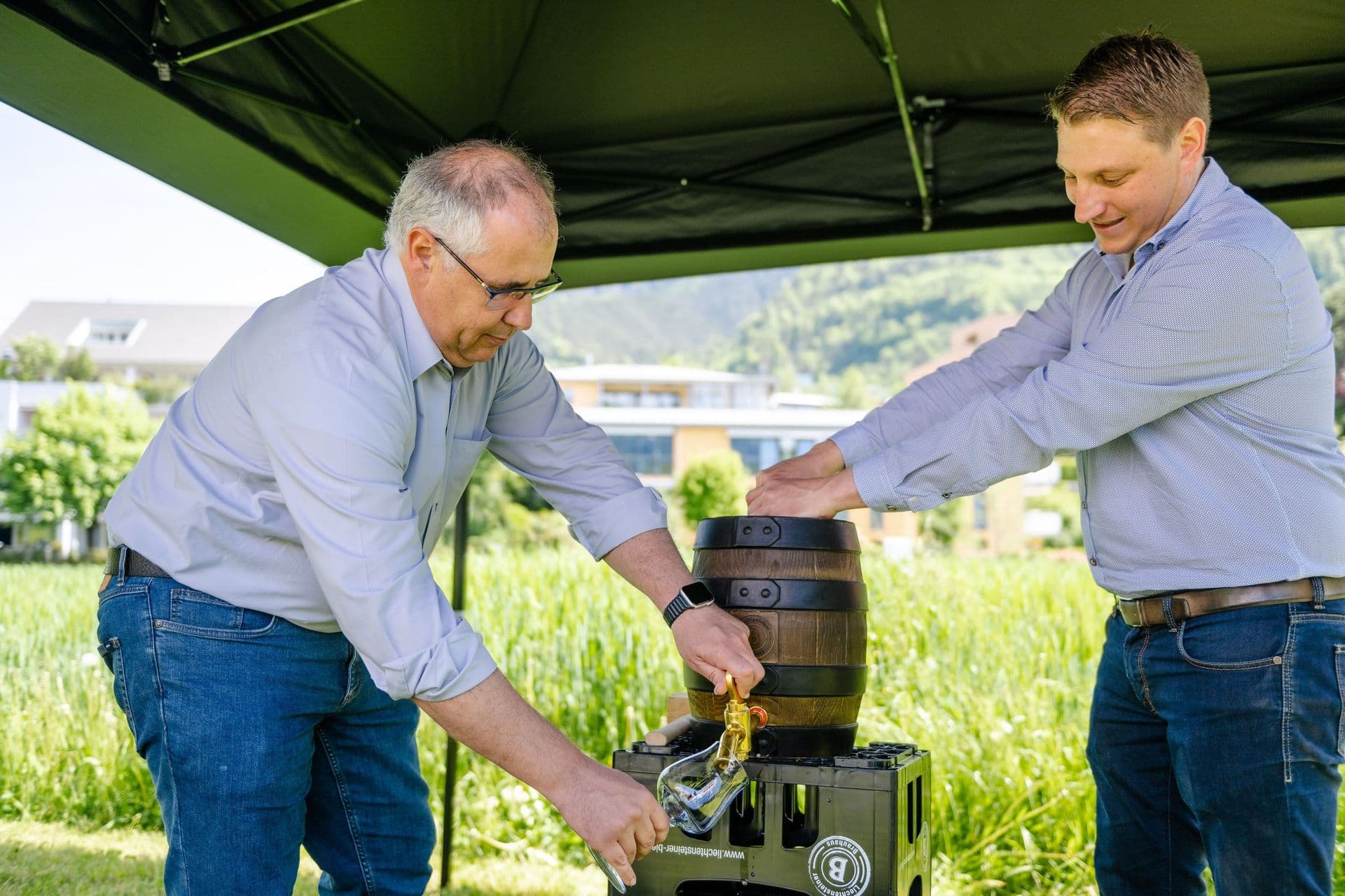 Alt-Bürgermeister Manfred Bischof und ein weiterer Mann zapfen im Freien unter einem Vordach Bier aus einem Holzfass, mit grünem Gras und Gebäuden im Hintergrund.