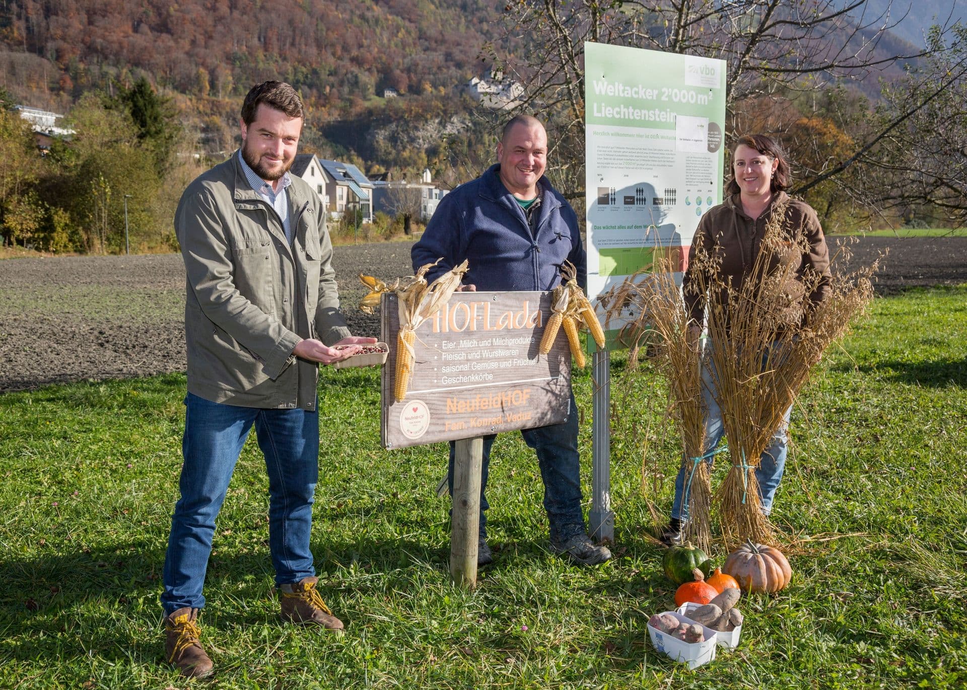 Drei Personen stehen im Freien neben einem Schild und Herbstprodukten, darunter Mais und Kürbisse, vor einer Kulisse aus Bäumen und Hügeln.