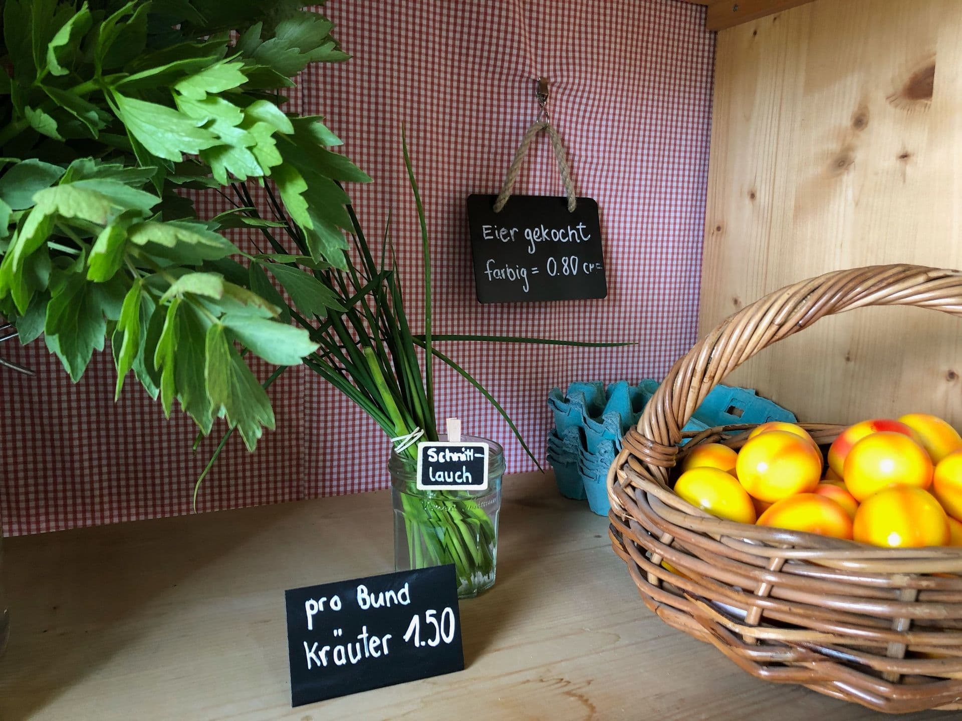 Frische Kräuter in einem Glas, Eier in einem Karton und ein Korb mit Pfirsichen in einem Holzregal mit Preisschildern.