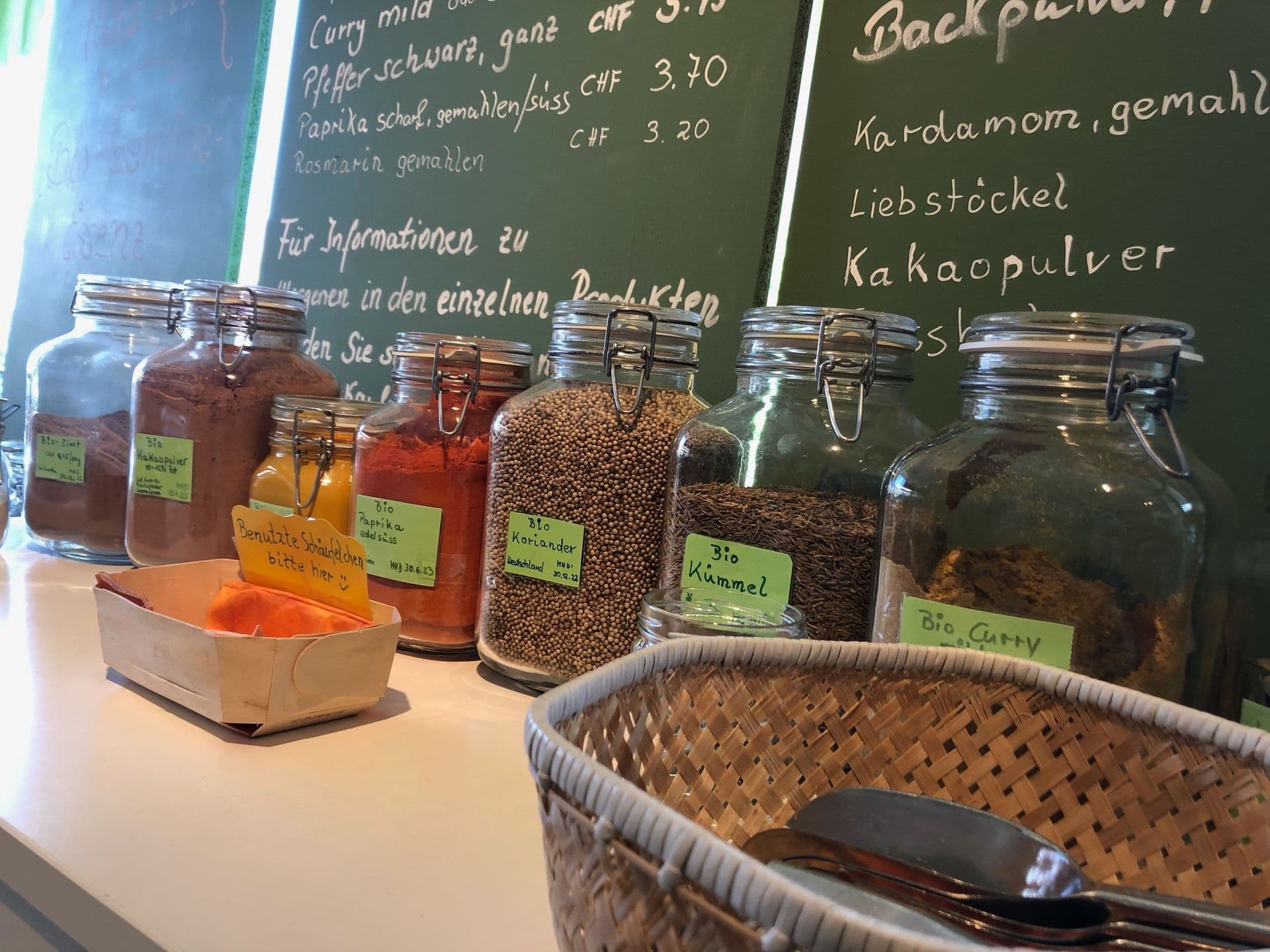 Gläser mit verschiedenen Gewürzen auf einer Theke, beschriftet in deutscher Sprache. Ein kleiner Korb mit Utensilien steht im Vordergrund.